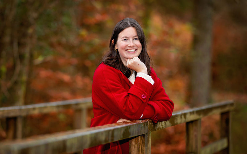 Smiling woman in a red coat resting her chin on her hand beside a wooden bridge railing in autumn woodland.