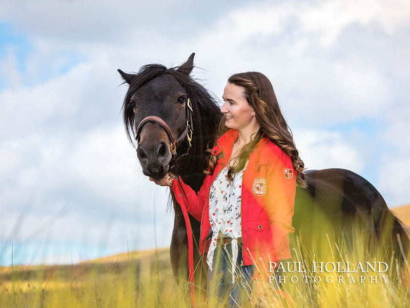 Woman in a red jacket standing beside a dark horse in tall grass beneath a cloudy sky.