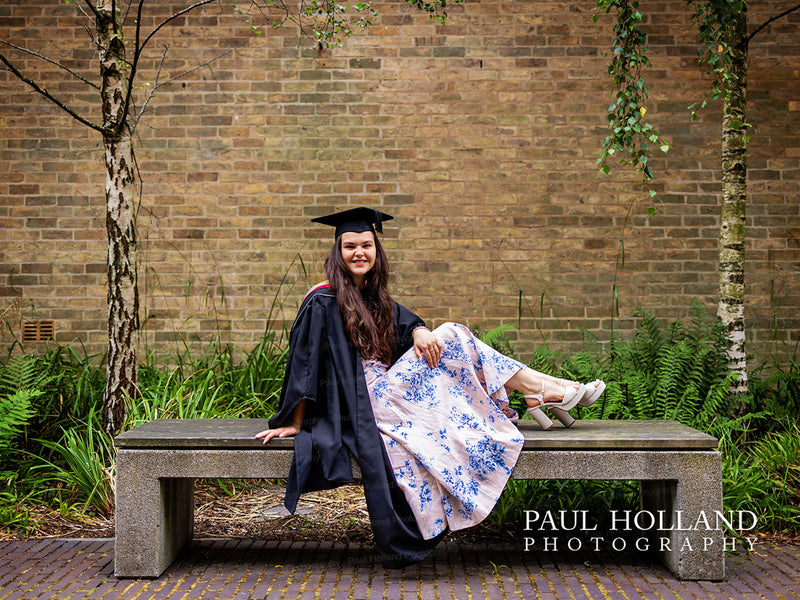 Graduate in cap and gown sitting on a bench with a brick wall and greenery in the background, photo by Paul Holland Photography.