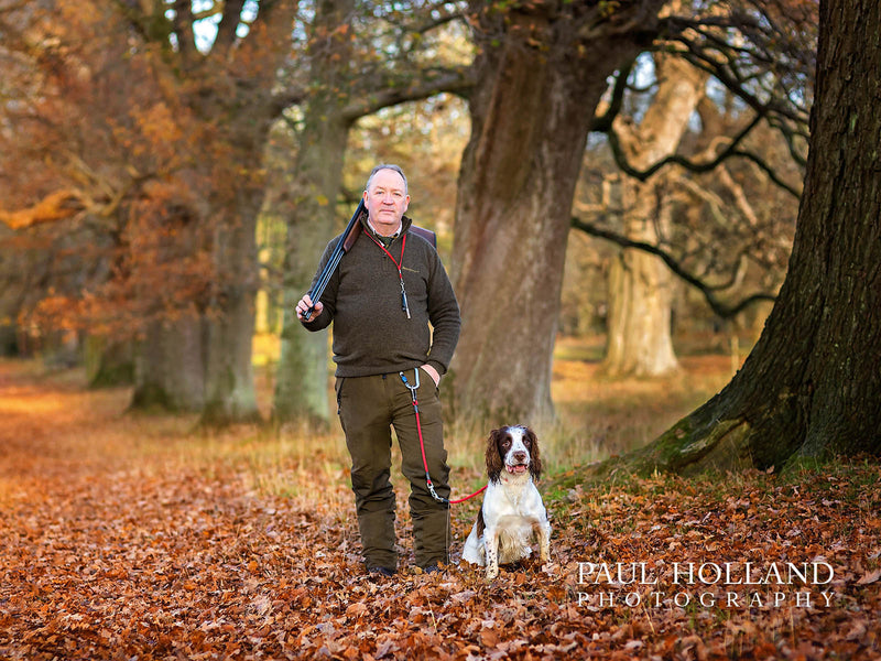 Man carrying a broken shotgun over his shoulder beside a liver and white spaniel dog amongst autumn woodland.